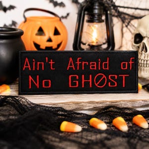 May include: A rectangular black embroidered patch featuring the red stitched text Ain't Afraid of No GHOST, with a red circle and diagonal line through the letter O in GHOST. The patch has a clean, finished border and is oriented horizontally.