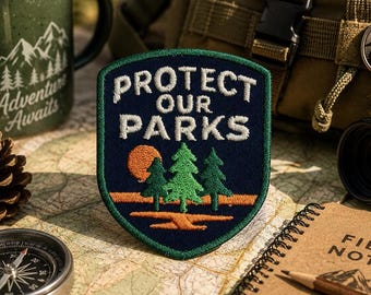 Puede incluir: Un parche bordado en forma de escudo con fondo azul oscuro y borde verde. El texto 'PROTECT OUR PARKS' está bordado en blanco sobre un gráfico de tres pinos verdes, un sol naranja y una línea de horizonte de color naranja.