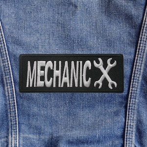 Può includere: Una toppa nera con la scritta "MECHANIC" ricamata in bianco, insieme a un disegno di chiavi incrociate. La toppa è cucita su un tessuto denim blu, probabilmente una giacca o una tuta.