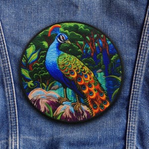 Puede incluir: Un parche bordado redondo con un pavo real. El pavo real tiene plumas azules, verdes y naranjas, sobre un fondo de exuberante follaje verde y una escena de bosque. El parche está sobre un fondo de mezclilla.