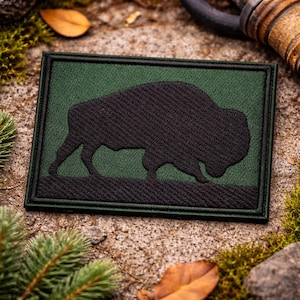 May include: A rectangular patch featuring a black bison silhouette on a green background, framed by a black border. The patch is made of textured fabric, and the bison is depicted in a walking pose. The patch is likely an emblem or a decorative item.