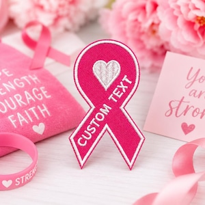May include: A hot pink embroidered ribbon patch featuring a white heart icon and the text CUSTOM TEXT in white thread. The patch has a white border and is displayed alongside a pink pouch with the words HOPE STRENGTH COURAGE FAITH, a pink silicone wristband with the word HOPE, and a pink card with the text You are strong.