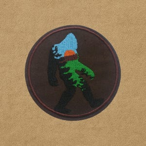 Peut inclure: Écusson brodé circulaire avec un motif de Bigfoot. Le patch présente une silhouette de Bigfoot en noir, vert, bleu, orange et marron, sur un fond beige. Le patch a une bordure marron et représente un Bigfoot en marche.