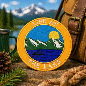 May include: Embroidered patch with a scenic lake design. The patch features a blue lake, green mountains, a yellow sun, and the text "LIFE AT THE LAKE" in white. The patch has a yellow border and is set against a natural background.