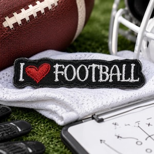 May include: A rectangular black embroidered patch featuring the white text I, a red heart symbol, and the white text FOOTBALL. The patch has a black stitched border and is positioned on a white mesh fabric surface with a football and helmet nearby.
