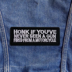 May include: A black rectangular patch with white embroidered text that reads "HONK IF YOU'VE NEVER SEEN A GUN FIRED FROM A MOTORCYCLE." The patch is sewn onto a blue denim jacket.