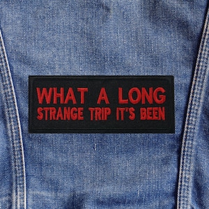 May include: A black rectangular patch with red embroidered text that reads "WHAT A LONG STRANGE TRIP IT'S BEEN." The patch is sewn onto a blue denim garment.