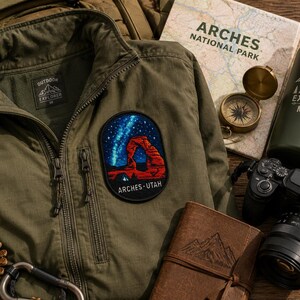 Op de afbeelding: Een olijfgroene jas met rits en een ovaal geborduurde patch die de rode rots Delicate Arch in Arches Utah onder een sterrenhemel afbeeldt. De patch bevat de tekst ARCHES UTAH in witte letters aan de onderkant.