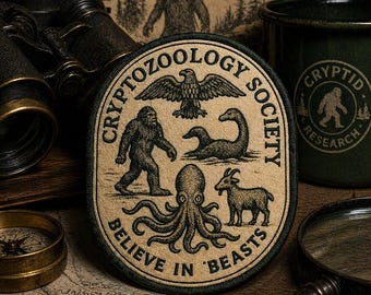 Peut inclure: Écusson de la Cryptid Zoology Society avec des illustrations de Bigfoot, d'un aigle, d'un serpent de mer, d'une pieuvre et d'une chèvre. L'écusson est beige avec une bordure verte et l'inscription "Believe in Beasts".
