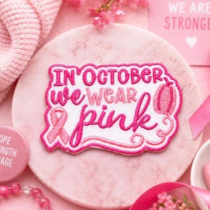 May include: A white embroidered patch with a pink border featuring the text "IN OCTOBER we wear pink" in pink thread. The design includes a pink awareness ribbon on the left and a small pink pumpkin icon on the right, with decorative swirls.