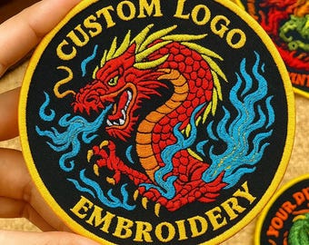 Könnte beinhalten: Runder, gestickter Aufnäher mit einem roten Drachen-Design, gelben und blauen Akzenten und den Worten "CUSTOM LOGO EMBROIDERY" in Gelb. Der Drache speit blaue Flammen. Der Aufnäher ist schwarz mit gelbem Rand.