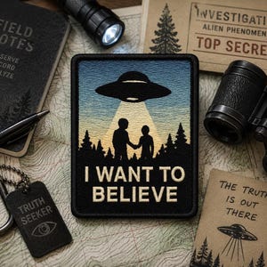 May include: A rectangular embroidered patch featuring a black UFO hovering over two silhouettes in a forest, illuminated by a bright yellow beam of light. The text I WANT TO BELIEVE is stitched in cream-coloured thread at the bottom of the patch.