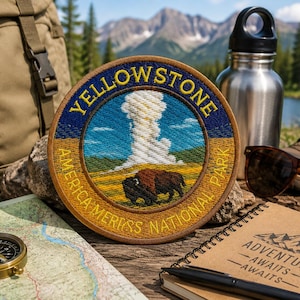 Op de afbeelding: Een ronde geborduurde patch met de tekst "YELLOWSTONE AMERICA'S NATIONAL PARK" rondom een afbeelding van een geiser en een bizon. De patch ligt op een houten oppervlak met een kompas, kaart en wandeluitrusting.