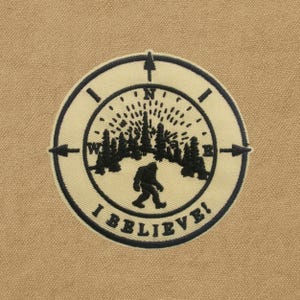 Peut inclure: Écusson circulaire avec un fond beige et un motif brodé noir et beige. Le motif représente une boussole, des arbres et une silhouette de Bigfoot, avec le texte "I BELIEVE!"