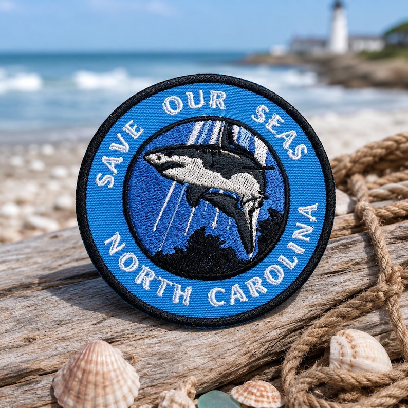 May include: A round, blue embroidered patch with the text "SAVE OUR SEAS NORTH CAROLINA". The patch features a black and white orca whale design in the center. The patch is on a piece of driftwood with seashells.