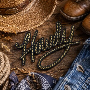 May include: A black embroidered patch featuring the word Howdy in a cursive script style. The lettering is outlined in black with a yellow rope-like texture pattern filling the interior of the letters. The patch is positioned on a dark wood surface.