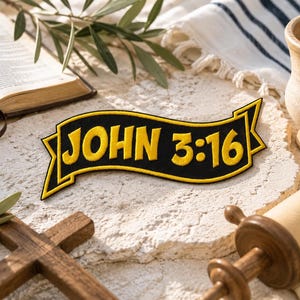 Puede incluir: Un parche bordado negro y amarillo con el texto "JOHN 3:16". El parche tiene forma de pancarta con un borde amarillo. La imagen también incluye una cruz de madera, un libro abierto y un rollo.