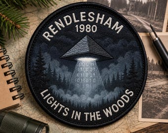 Può includere: Una toppa circolare ricamata che raffigura una scena di bosco scuro con un velivolo triangolare in volo stazionario. Il testo recita RENDLESHAM 1980 e LIGHTS IN THE WOODS. Un raggio di luce con codice binario scende sugli alberi.