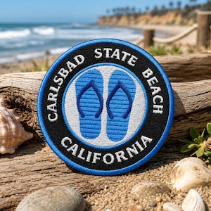 May include: A circular embroidered patch featuring a black border with the white text Carlsbad State Beach California. The centre displays two light blue flip-flops on a white background, all enclosed by a thin blue stitched edge.