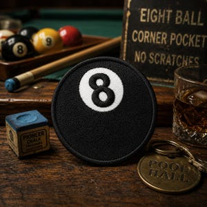 May include: A black and white embroidered patch of an eight ball, with the number 8 in white. The image includes a pool cue, chalk, a glass of whiskey, and a sign that reads "EIGHT BALL CORNER POCKET NO SCRATCHES". A brass key chain reads "POOL HALL".