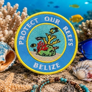May include: A circular embroidered patch with a light blue border and a yellow edge. The center features a coral reef illustration with the text "PROTECT OUR REEFS" arched above and "BELIZE" printed in white lettering at the bottom of the patch.