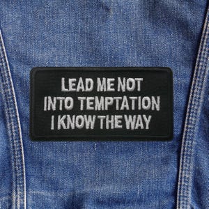 May include: A black rectangular patch with white embroidered text that reads "LEAD ME NOT INTO TEMPTATION I KNOW THE WAY." The patch is affixed to a blue denim garment, with visible stitching and seams.
