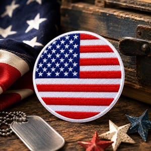 May include: A circular embroidered patch featuring the United States flag with a blue field of white stars on the left and horizontal red and white stripes on the right, finished with a white stitched border around the entire circular edge.