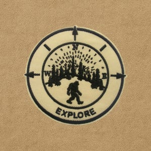 May include: A circular patch with a compass design, featuring a silhouette of a Bigfoot figure walking towards a forest. The word "EXPLORE" is printed in a bold, black font at the bottom. The patch is beige with black embroidery.