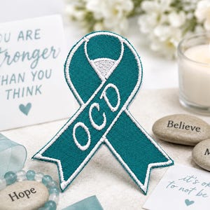 May include: A teal OCD awareness ribbon with white embroidered lettering. The image includes a card with the text "You are stronger than you think", a lit candle, and inspirational stones with the words "Believe" and "You Matter".