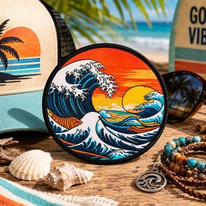 May include: A circular embroidered patch featuring a stylized ocean wave in shades of blue and white against a vibrant orange and yellow sunset background, with a black stitched border, displayed alongside a hat, sunglasses, and beaded bracelets.