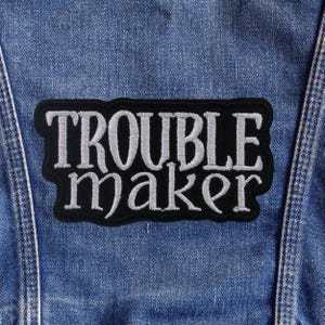 Puede incluir: Un parche bordado en blanco y negro con las palabras "TROUBLE MAKER" en una fuente estilizada. El parche es rectangular con un borde negro y está adherido a una chaqueta vaquera azul.