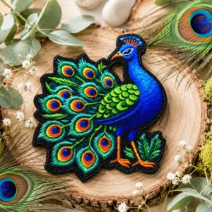 Puede incluir: Parche de pavo real bordado con detalles vibrantes en azul, verde y naranja. El parche presenta un diseño detallado de un pavo real con las plumas de la cola extendidas. El parche está sobre una superficie de madera, rodeado de vegetación y plumas de pavo real.