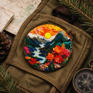 Puede incluir: Parche bordado redondo que representa un paisaje de montaña con un río, una puesta de sol y flores coloridas. El parche está ricamente detallado con varios colores. Descansa sobre un fondo de lona verde, acompañado de una brújula y un mapa.