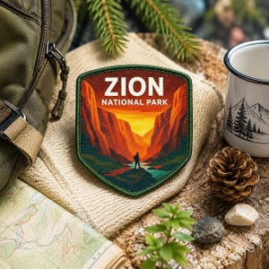 May include: A Zion National Park patch with a landscape design featuring a hiker, canyon walls, and a sunset. The patch is green, orange, and brown. A map, mug, and backpack are in the background.