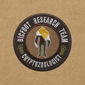 Peut inclure: Écusson brodé circulaire avec une illustration de Bigfoot en marron, vert et jaune. Le texte "BIGFOOT RESEARCH TEAM CRYPTOZOOLOGIST" entoure le bord. Comprend deux empreintes de pas de Bigfoot vertes. Fond beige.