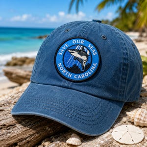 May include: A blue baseball cap with a circular patch that reads "Save Our Seas North Carolina." The patch features a shark graphic. The hat is sitting on a piece of driftwood on a sandy beach with the ocean in the background.