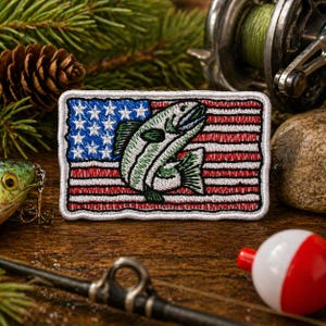 May include: Embroidered patch featuring a leaping fish over an American flag design. The patch has a white border, blue stars, and red and white stripes. Fishing gear, including a reel, lure, and bobber, surrounds the patch on a wooden surface.