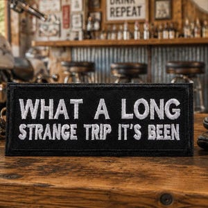 May include: A black rectangular patch with white embroidered text that reads "WHAT A LONG STRANGE TRIP IT'S BEEN." The patch is on a wooden surface, with a motorcycle and bar in the background. Leather gloves are visible.