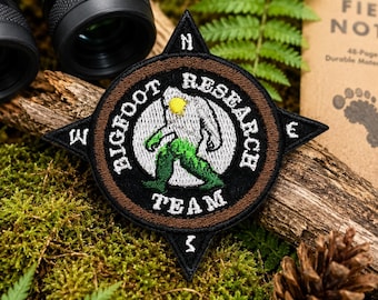 May include: A circular embroidered patch shaped like a compass with four points. The black border features the text BIGFOOT RESEARCH TEAM. The center depicts a white creature with green legs and a yellow head. The patch has brown and black accents.