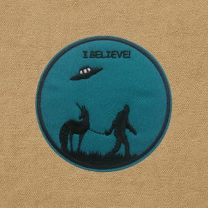 May include: Embroidered patch with a teal background and a black border. The patch features a UFO, a unicorn, and a Bigfoot-like figure walking with the unicorn on a leash. The text "I BELIEVE!" is arched above the UFO.