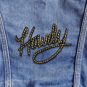 Könnte beinhalten: Nahaufnahme einer blauen Jeansjacke mit einem gold-schwarzen gestickten Aufnäher. Der Aufnäher zeigt das Wort "Howdy" in einer stilisierten, seilartigen Schriftart und verleiht dem Kleidungsstück einen Hauch von Western-Flair.