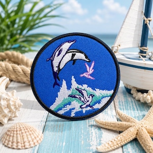 May include: Round blue embroidered patch featuring two dolphins leaping above waves, with a seagull and a small bird. The patch has a black border. The design is reminiscent of the ocean and marine life.