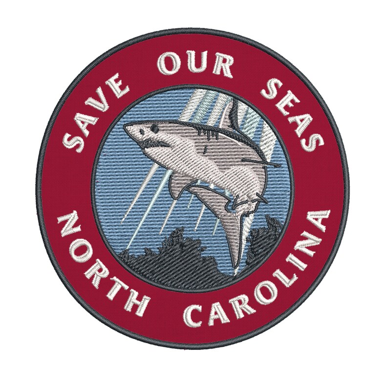 May include: Embroidered patch with a shark leaping from the ocean. The patch is circular with a red border and the text "SAVE OUR SEAS NORTH CAROLINA" in white. The shark is gray and white.