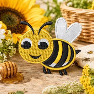 May include: An embroidered bee patch featuring a yellow and black striped body, a round yellow head with large white eyes, black pupils, and two white wings. The bee has two black antennae and four small black legs extending from the bottom.