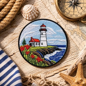 May include: Embroidered patch featuring a lighthouse scene with a red-roofed building, white tower, and red flowers. The patch is round with a black border, set against a blue sky and ocean. The design evokes a nautical theme.