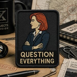 May include: A rectangular embroidered patch with a black border featuring a stylised illustration of a person with shoulder-length red hair wearing a dark blue blazer over a white shirt. The text "QUESTION EVERYTHING" is embroidered in beige.