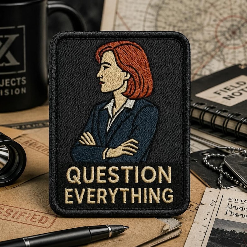 May include: A rectangular embroidered patch with a black border featuring a stylized illustration of a person with shoulder-length red hair wearing a dark blue blazer over a white shirt. The text "QUESTION EVERYTHING" is embroidered in beige.
