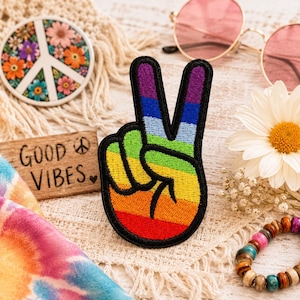 May include: A colorful embroidered patch in the shape of a peace sign hand gesture, featuring a rainbow design with a black border. The image also includes a wooden sign that says "GOOD VIBES", a peace sign coaster, and a pair of pink sunglasses.