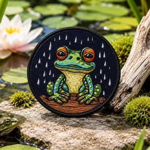 Pode incluir: Um emblema bordado circular apresentando um sapo verde com grandes olhos laranja sentado num tronco castanho contra um fundo azul escuro com riscas de chuva verticais brancas. O sapo tem pele amarela e verde manchada.
