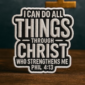 May include: Embroidered patch with the text "I CAN DO ALL THINGS THROUGH CHRIST WHO STRENGTHENS ME PHIL 4:13" in white thread on a black background. A gold cross is in the background. The patch is on a wooden surface.